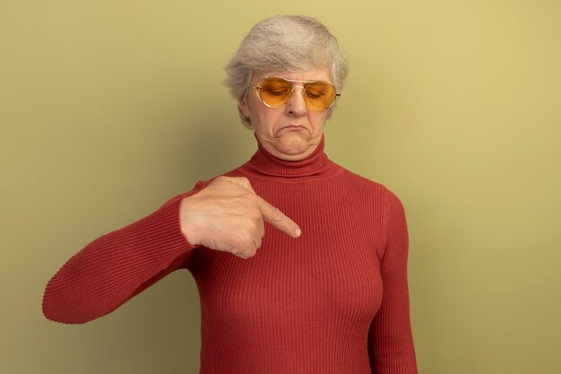 Oude vrouw met een rode coltrui en een zonnebril die naar haar borst kijkt en wijst