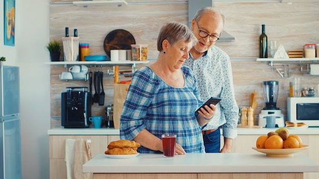 Ouder stel tijdens videochat met familie die smartphone in de keuken gebruikt. Grootouders online gesprek. Ouderen met moderne technologie op pensioengerechtigde leeftijd die mobiele apps gebruiken