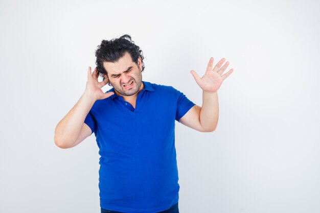 Oudere man die oor met vinger inplugt, stopgebaar in blauw t-shirt toont en geïrriteerd kijkt. vooraanzicht.