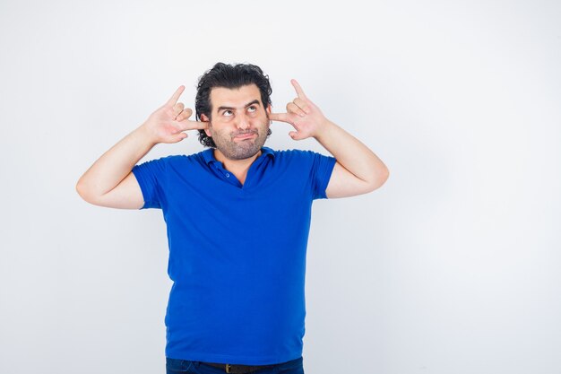 Oudere man in blauw t-shirt oren met vingers inpluggen, lippen gebogen en peinzend, vooraanzicht op zoek.