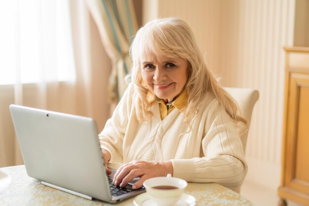 Oudere mooie vrouw achter een computer drinkt een kopje thee en geniet van haar vrije tijd