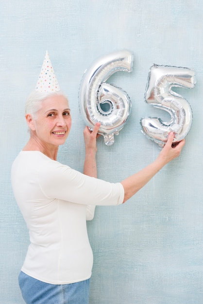 Oudere mooie vrouw die het metaalnummer van de folieballon op haar verjaardag houden