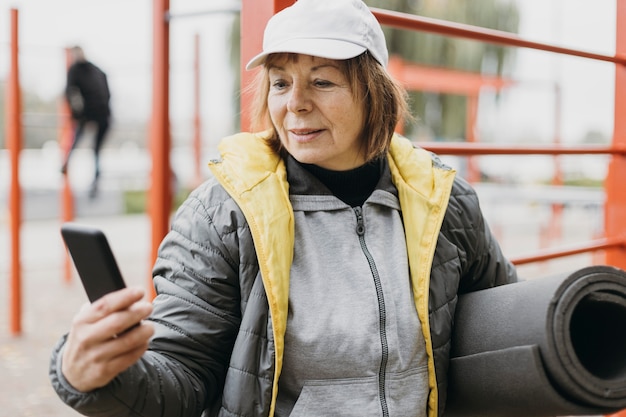 Oudere vrouw buitenshuis trainen terwijl ze smartphone en mat vasthoudt