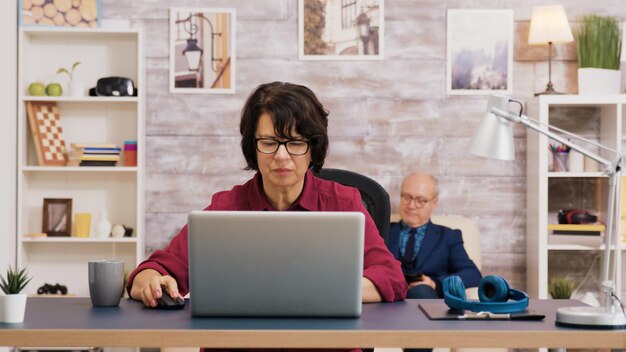 Oudere vrouw die een slokje koffie neemt tijdens het browsen op de laptop. Oude man ontspannen op de bank op de achtergrond.