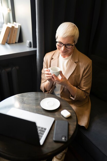 Oudere vrouw die koffie drinkt in een café terwijl ze naar haar laptop kijkt