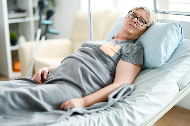 Oudere vrouw die op een bed ligt in een verpleeghuis bedekt met een deken.