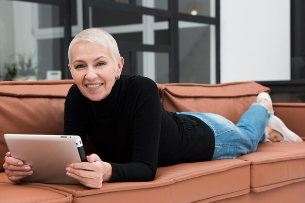 Oudere vrouw die terwijl het zitten op bank en het houden van tablet glimlacht