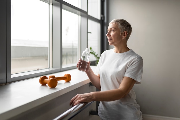 Oudere vrouw die water drinkt in de sportschool terwijl ze een pauze neemt