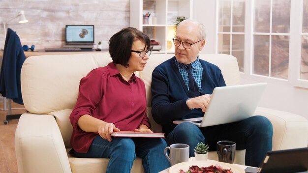 Ouderen echtpaar met behulp van laptop zittend op de bank in de woonkamer. Bejaarde vrouw die een slokje koffie neemt.