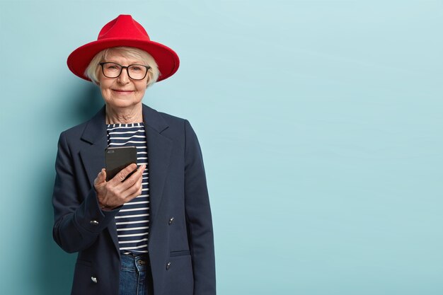 Ouderen en technologieconcept. Senior vrouw blijft altijd verbonden, chat via app, stuurt berichten, draagt rode hoofddeksel, gestreepte trui met formele jas, geïsoleerd over blauwe muur