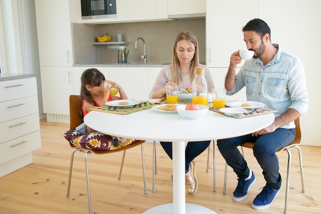 Ouders en dochter samen ontbijten, koffie en sinaasappelsap drinken, zittend aan eettafel met fruit en koekjes.