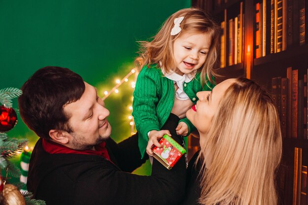 Ouders met een klein blond meisje poseren voor een kerstboom in de kamer