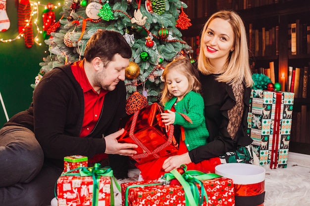 Ouders met een klein blond meisje poseren voor een kerstboom in de kamer