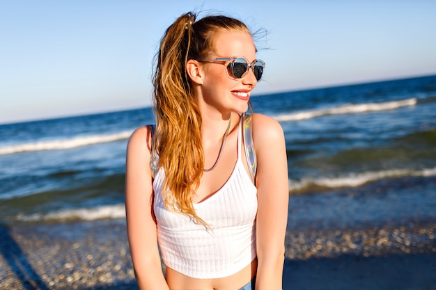 Outdoor lifestyle portret van grappig gelukkig meisje dat alleen naar de oceaan reist, glimlachend en geniet van zee strandtijd, gelukkige positieve emoties, gespiegelde zonnebril, witte crop top en rugzak, vreugde, beweging.