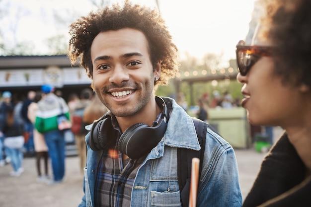 Gratis foto outdoor portret van charmante afro-amerikaanse man wandelen met vriend in park, denim kleding en koptelefoon dragen over nek,