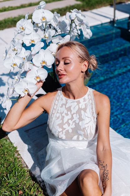 Outdoor Portret van een vrouw in een witte trouwjurk, zittend in de buurt van blauwe zwembad met bloemen