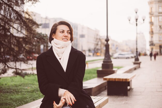 Outdoor Portret van Kaukasische vrouw, gekleed in zwarte winterjas en sjaal zittend op de Bank, stad op achtergrond