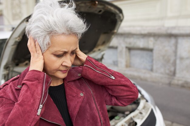 Outdoor Portret van ongelukkig gestresste vrouwelijke gepensioneerde m / v met kort grijs haar voor oren, gefrustreerd omdat haar auto kapot is
