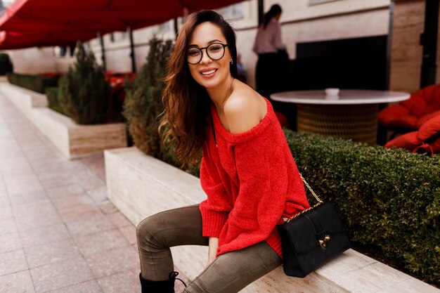 Outdoor Portret van succesvolle lachende brunette vrouw in trendy rode gebreide trui zitten en genieten van Europese feestdagen. Elegante zwarte tas.