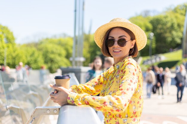 Outdoor Portret van vrouw in gele zomerjurk en hoed met kopje koffie genieten van zon, staat op brug met prachtig uitzicht op de stad