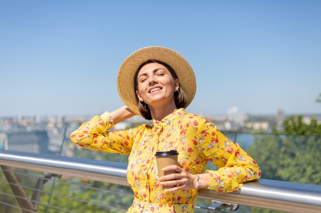 Outdoor portret van vrouw in gele zomerjurk en hoed met kopje koffie genieten van zon, staat op brug met prachtig uitzicht op de stad