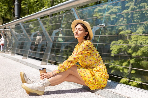 Outdoor Portret van vrouw in gele zomerjurk en hoed met kopje koffie genieten van zon