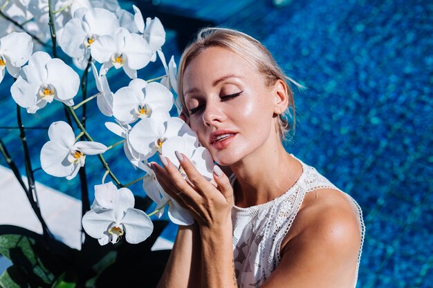 Outdoor Portret van vrouw in witte trouwjurk zitten in de buurt van blauwe zwembad met bloemen orchidee