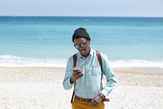 Outdoor shot van Afro-Amerikaanse jonge man met rugzak, hoed, tinten en stijlvolle kleding aan het wachten op zijn date op kiezelstrand, met behulp van 3g of 4g internetverbinding op touchscreen mobiele telefoon