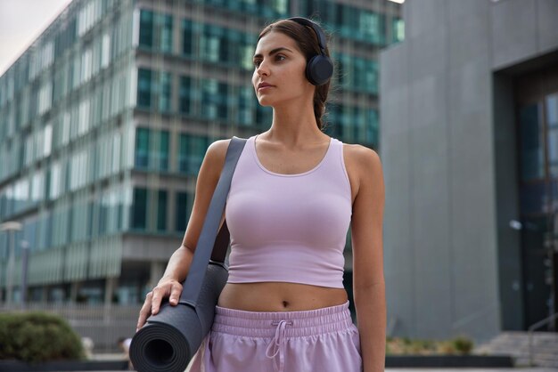 Outdoor shot van peinzende sportieve vrouw gekleed in bijgesneden top en korte broek heeft een goede fysieke vorm draagt opgerolde karemat luistert muziek via draadloze koptelefoon poses tegen modern stadsgebouw