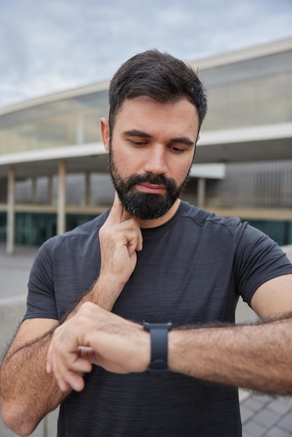 Outdoor shot van serieuze bebaarde volwassen man controleert hartslag na training kijkt smartwatch draagt casual zwarte t-shirt poses tegen wazige monitoren hartslag maakt gebruik van sporttechnologie