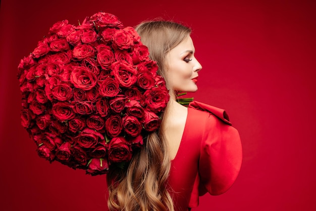 Over schouder studio portret van prachtige jonge brunette met heldere lippen in rode jurk met groot boeket rode rozen en glimlachend in de camera over rode achtergrond isoleren op rood