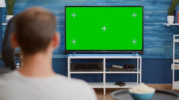 Over schouder zicht van man kijken naar film op tv met groen scherm ontspannen met kom popcorn zittend op de bank. Achteraanzicht van persoon ontspannen op de bank voor televisiemodel met chroma key disp