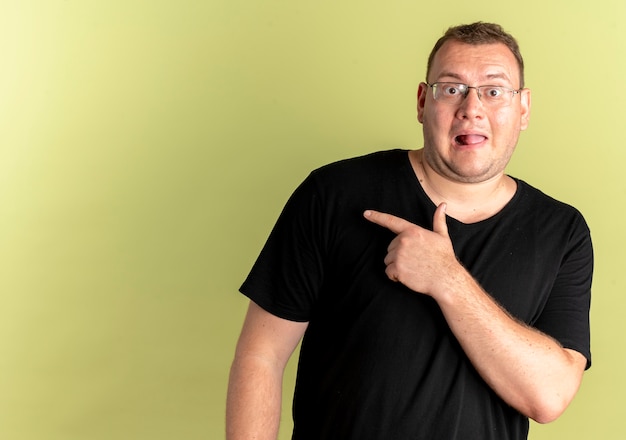 Gratis foto overgewicht man in bril dragen zwart t-shirt blij en verrast wijzend met de vinger naar de zijkant staande over lichte muur