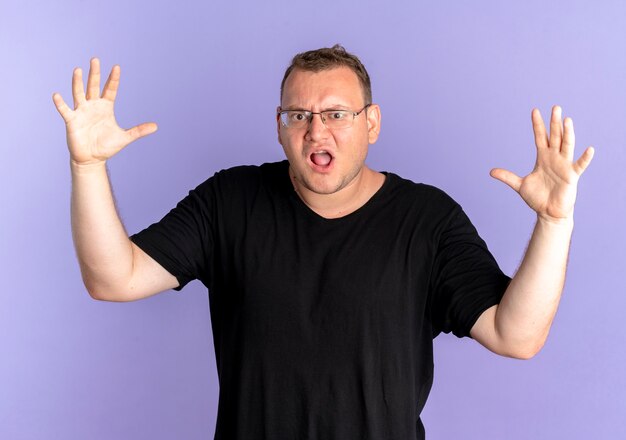 Overgewicht man in bril met zwart t-shirt gefrustreerd wild gaan schreeuwen met een boos gezicht en opgeheven armen over blauw