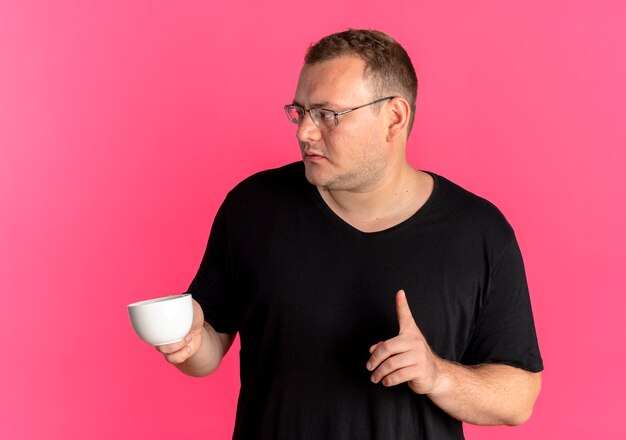 Overgewicht man in glazen dragen zwart t-shirt met koffiekopje wijsvinger tonen over roze