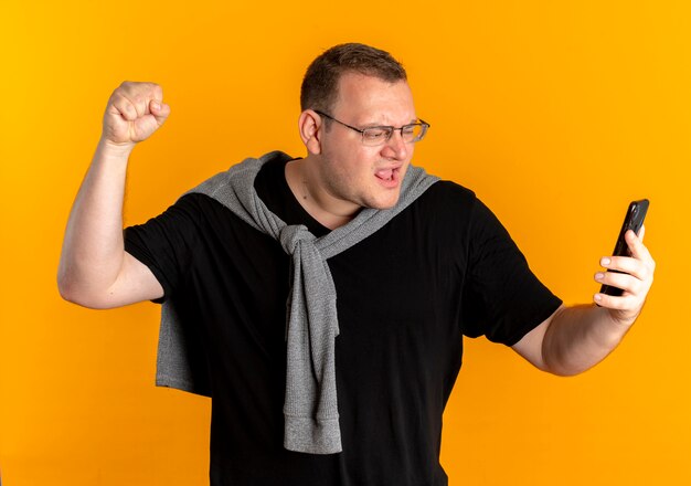 Overgewicht man in glazen dragen zwart t-shirt met smartphone clanching vuist vreugde zijn succes over oranje