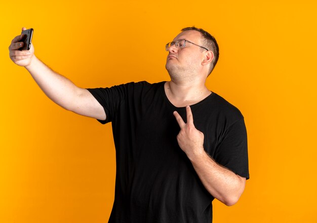 Overgewicht man in glazen dragen zwarte t-shirt met smartphone maken selfie weergegeven: overwinningsteken staande over oranje muur