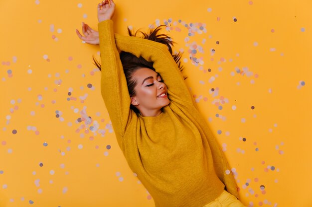 Overhead schot van zorgeloos meisje liggend op confetti met glimlach. Binnenfoto van elegant donkerbruin model in gele sweater.