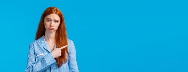 Gratis foto overstuur dwaas en timide schattig tienermeisje met rood lang haar dat mokkend en fronsend nachtkleding draagt