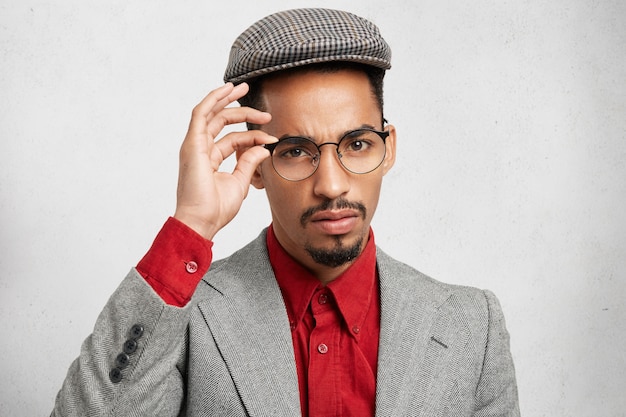 Overtuigde zelfverzekerde zwarte machoman draagt trendy bril, pet en jas,