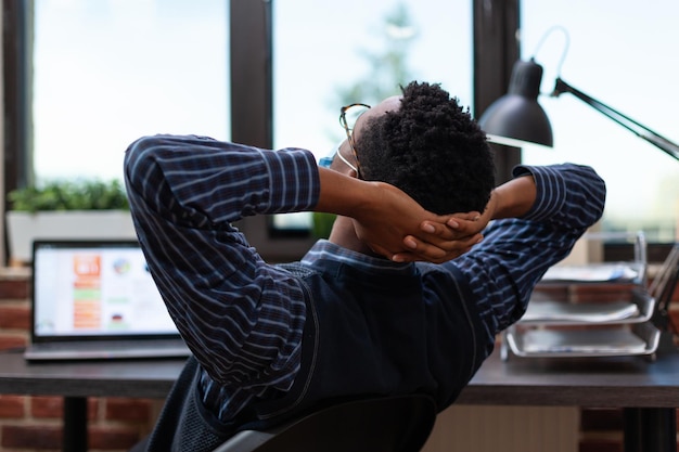 Overwerkte werknemer die een covid-beschermingsmasker draagt en een dutje doet op het werk in de bureaustoel. Vermoeide startende ondernemer die zich uitstrekt en een pauze neemt van het werken op een laptop met grafieken.