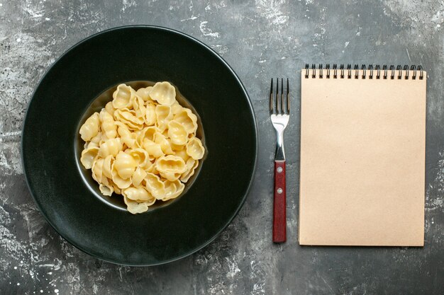 Overzichtsmening van heerlijke conchiglie op een zwarte plaat en een mes naast notitieboekje op grijze achtergrond