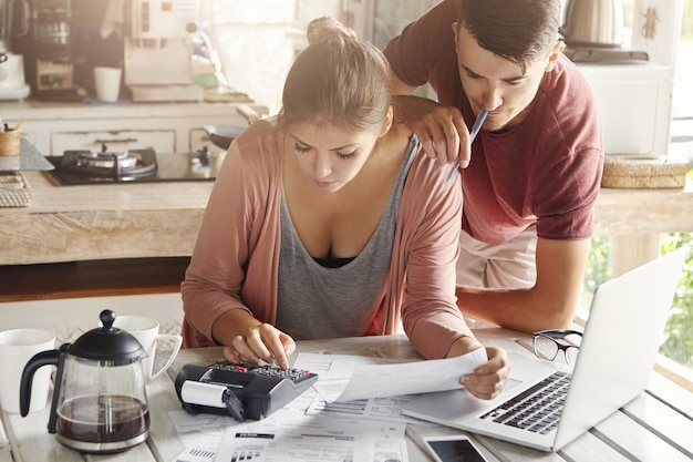 Paar geconfronteerd met financiële stress. Het jonge wijfje kleedde terloops het plannen van gezinsbudget in keuken, gebruikend calculator. Haar man houdt pen vast en staat naast haar