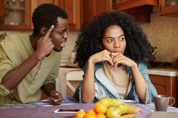 Paar geschil. Geërgerd mooie donkere vrouw zittend aan de keukentafel, negeert geschreeuw en beledigingen van haar gekke woedende echtgenoot die tegen haar schreeuwt, terwijl ze zijn vinger vasthoudt aan zijn slaap