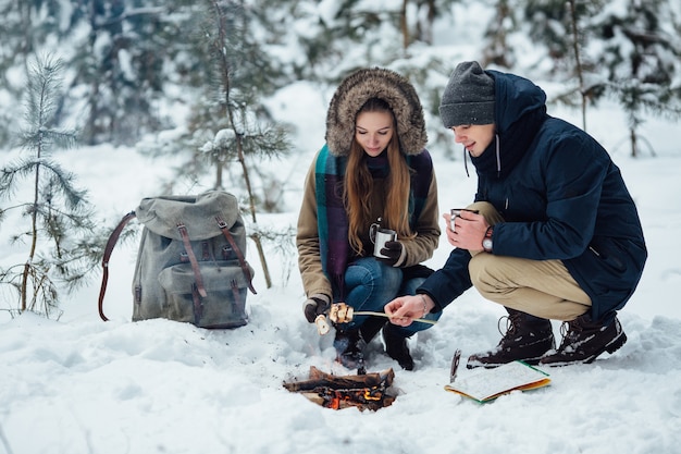 Paar jonge reizigers die heemst roosteren over vuur in sneeuw de winterbos