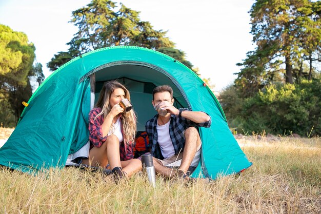 Paar thee drinken en kijken naar natuurlandschap. Aantrekkelijke blanke toeristen ontspannen in tent, genieten van landschap en zittend op het gazon. Backpacken toerisme, avontuur en zomervakantie concept