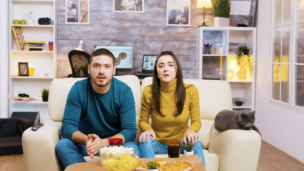 Paar zittend op de bank juichen tijdens het kijken naar sport op tv en het eten van junkfood met hun kat naast hen.