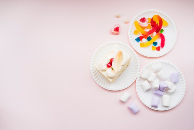 Papieren borden met marshmallows; gelei snoepjes en plak cake op roze achtergrond