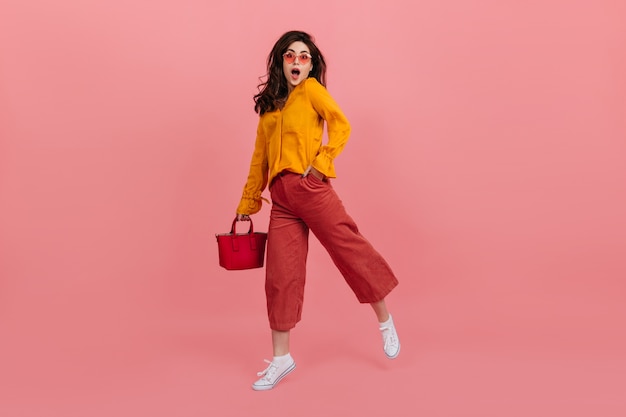 Parmantig meisje in een stijlvolle bril staart met verbazing, lopend op roze muur. Brunette in culottes en oranje blouse poseren met rode handtas.