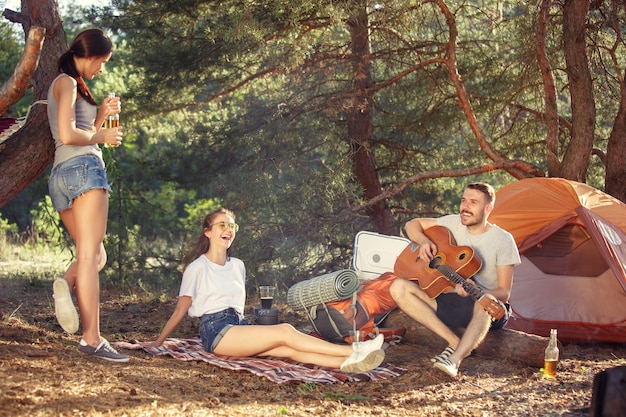 Partij, kamperen van mannen en vrouwengroep bij bos. Ze ontspanden zich en zongen een liedje tegen het groene gras. Het concept vakantie, zomer, avontuur, levensstijl, picknick